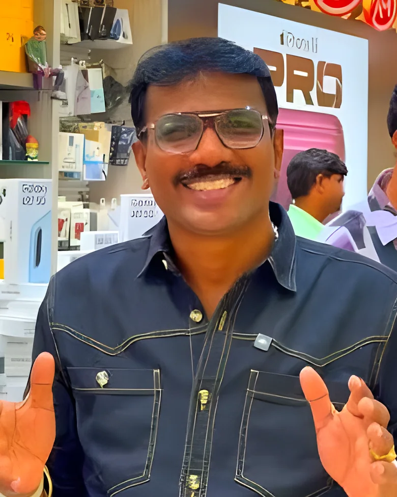 Eswar Datt, in a dark shirt with his hands raised mid-sentence, smiling in front of the display shelves inside the Arudra Mobiles shop.