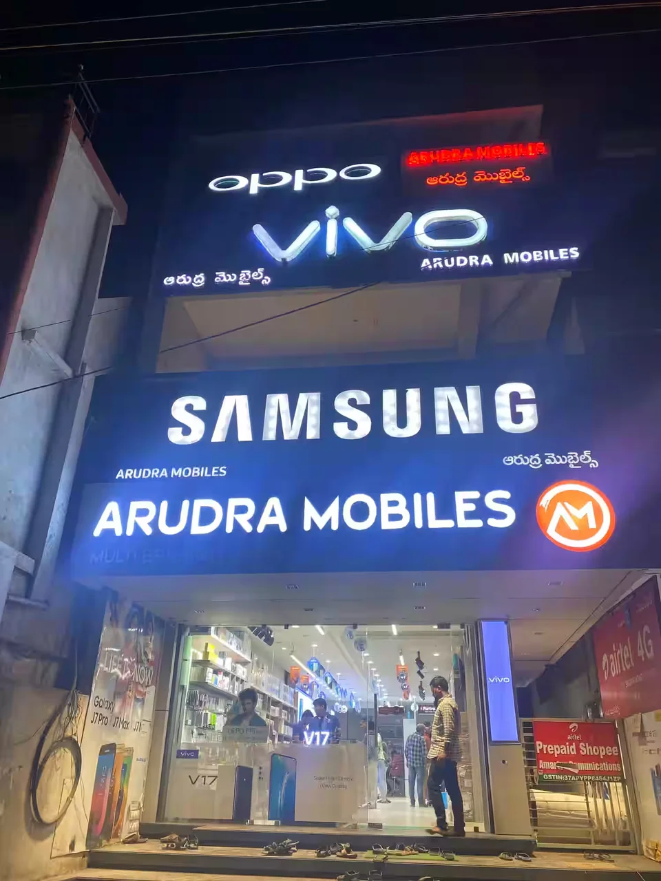The Arudra Mobiles shopfront at night: the lit Samsung signboard with the Arudra Mobiles name and its orange circular mark below it, and the open shop beneath showing display counters, shelves and customers inside.